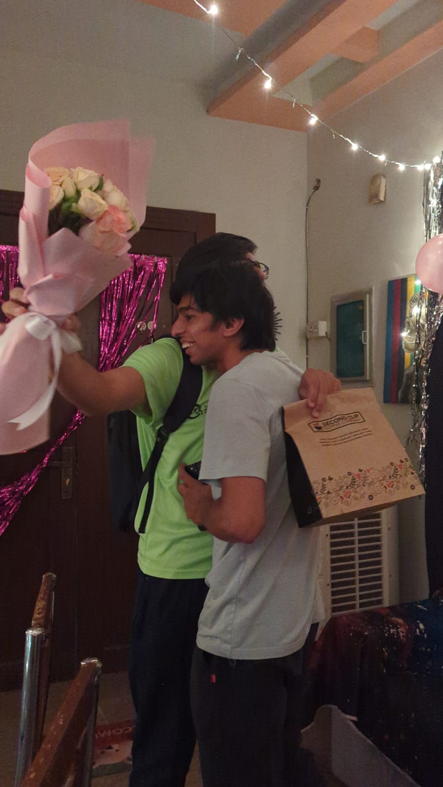 Walking in through a doorway holding a bouquet of pale roses and a Second Cup bag, fairy lights and pink streamers overhead.